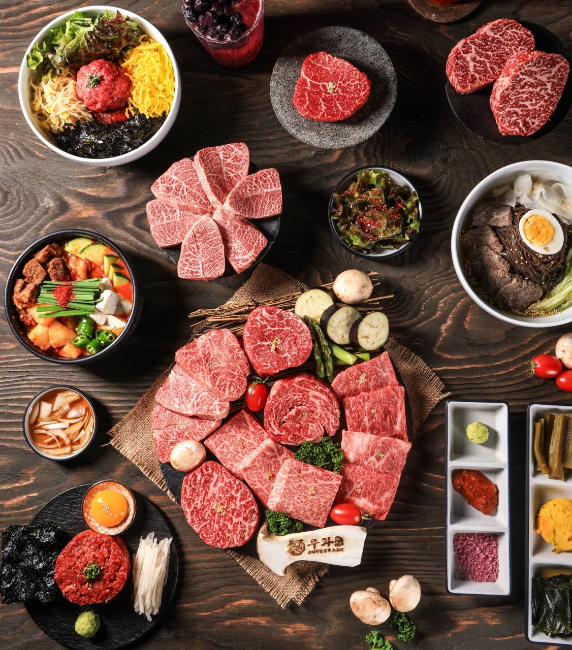 위에서 내려다본 1++ 한우 한 상 — 마블링이 선명한 생고기를 꽃 모양으로 말아 담은 접시들, 육회와 달걀노른자, 한우 냉면, 버섯과 채소, 세 칸 양념 접시가 어두운 나무 상판에 함께 놓여 있다