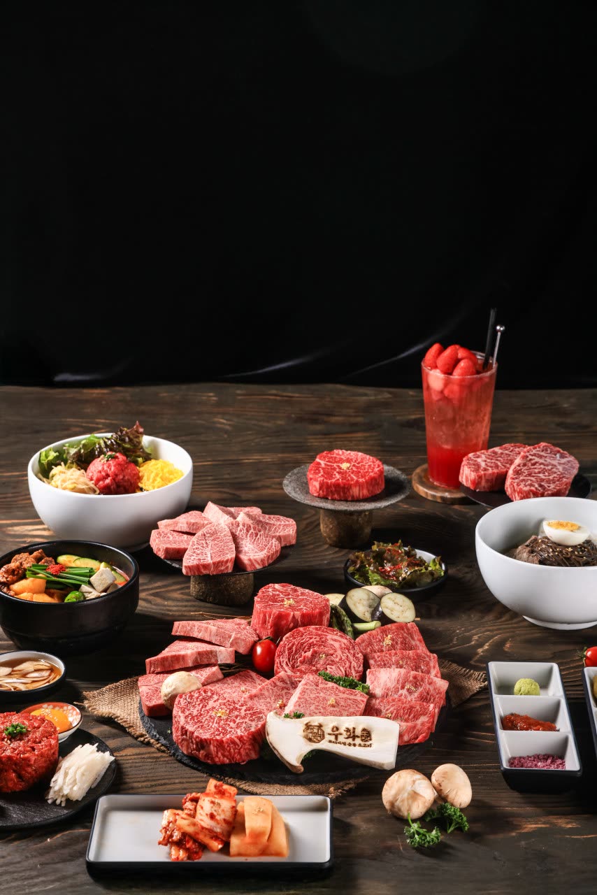 어두운 나무 식탁에 차려진 한 상 — 생고기 접시 여러 개, 육회, 육회 비빔밥, 반찬 종지들, 구운 채소, 그리고 딸기가 올라간 붉은 에이드 한 잔
