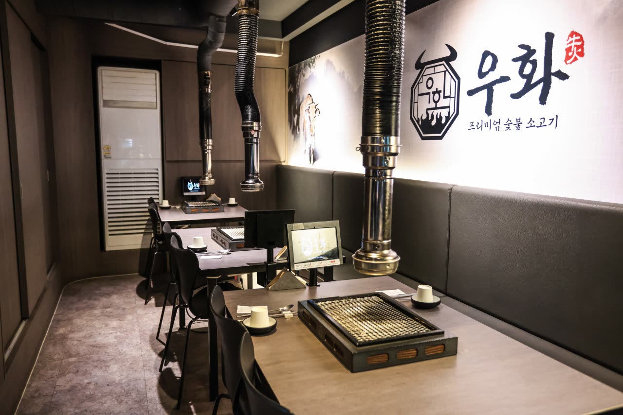 수묵화 벽과 우화 로고가 걸린 안쪽 자리. 숯불 그릴이 놓인 테이블과 등받이 없는 벤치, 태블릿이 함께 보인다
