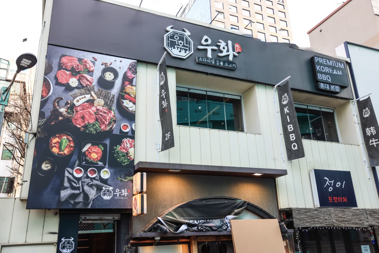 낮에 본 우화 홍대본점 건물 2층 외관. 검은 외벽에 고기 요리 사진이 크게 붙어 있고, 오른쪽에 'PREMIUM KOREAN BBQ 홍대 본점' 간판과 '韓牛', '2F' 배너가 걸려 있다