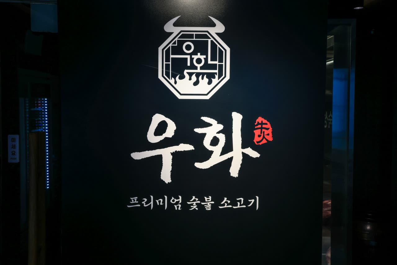 어둠 속에서 빛나는 우화 홍대본점 간판 — 소머리를 감싼 팔각 엠블럼 아래 붓글씨 '우화'와 붉은 낙관, 그 밑에 '프리미엄 숯불 소고기'라고 적힌 홍대 한우 맛집의 밤 간판