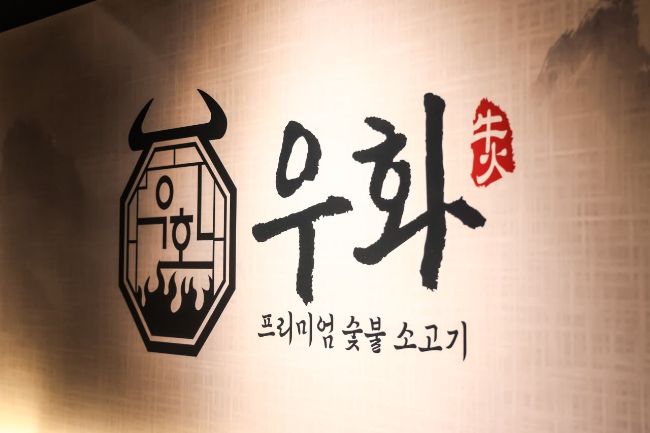 벽에 크게 그려진 우화 로고 — 팔각 엠블럼 안의 소머리와 불꽃, 붓글씨 '우화'와 붉은 낙관, 그 아래 '프리미엄 숯불 소고기'